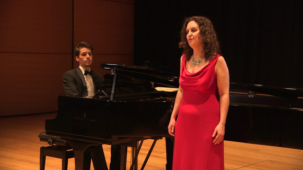 Soprano Ruth Hopkins with curly hair wearing a long red dress stands next to pianist Duncan Appleby.